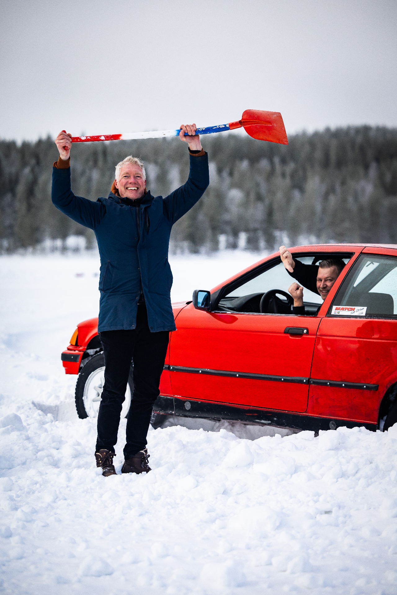 Deelnemer met schep naast rode driftauto in de sneeuw